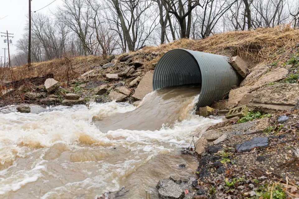Large water pipe with brown water pouring out into dirt ditch.